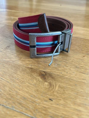 Randigt rött bälte med metallspänne - Snyggt Original Pinguin bälte i rött med blå och vita ränder. Bältet har ett robust metallspänne och detaljer i brunt skinn. Perfekt för att ge din outfit en färgklick och en sportig touch.