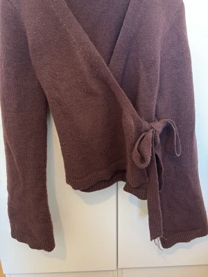 Brun omlottstickad tröja - Mysig brun stickad tröja i omlottmodell med knytband i sidan. Tröjan har långa ärmar och ribbade muddar vid ärmslut och nederkant. Perfekt för lager på lager och ger en avslappnad men ändå stilren look.