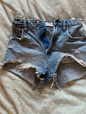 Blå slitna jeansshorts med fransar - Säljer ett par ljusblå jeansshorts från ZARA med råa kanter och slitningar. Shortsen har fem fickor, knapp och dragkedja framtill samt fransiga detaljer längs benen. Perfekta för sommaren och ger en cool, avslappnad vibe.
