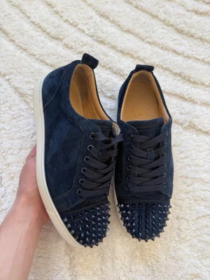 Mörkblå sneakers från Louboutin - Säljer ett par mörkblå sneakers från Louboutin med klassisk röd sula och coola nitar på hälen. Skorna är i mocka och har vita sulor. Perfekta för dig som vill sticka ut med en lyxig och edgy stil. Självklart äkta, kan ta bild på det som önskas.