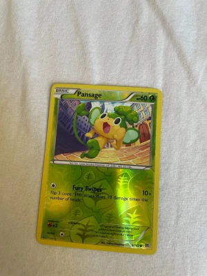 Pokémonkort Pansage holo - Säljer ett holografiskt Pokémonkort med Pansage. Kortet har guldkant och motivet visar Pansage i en stadsmiljö. Text och detaljer på engelska, med gröna och gula färger. Perfekt för samlare eller Pokémonfans.
