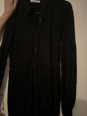 Svart spetsklänning från NA-KD - Svart långärmad klänning från NA-KD i spetsmaterial med knappar framtill. Klänningen har en elegant och figurnära passform med fina spetsdetaljer som ger en lyxig känsla. Perfekt för dig som vill ha en stilren och trendig look.