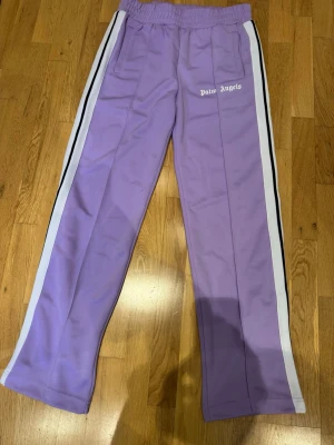 Lila mjukisbyxor  - Snygga lila mjukisbyxor  Palm Angels med vita och svarta ränder längs sidorna. Byxorna har resår i midjan, raka ben och två sidofickor. Palm Angels-logga tryckt på vänster sida. Perfekta för en chill och trendig look.