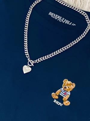 Silverfärgat halsband med hjärthänge - Säljer ett chunky halsband i silverfärg rostfritt stål med ett stilrent hjärthänge. Halsbandet har en bred kedja och ett klassiskt T-lås framtill. Perfekt accessoar för att lyfta din outfit med en trendig touch.