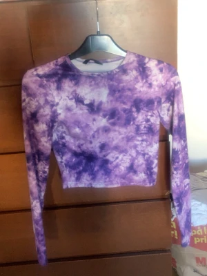 Lila tie-dye croppad långärmad topp - Trendig croppad långärmad topp i lila och vit tie-dye. Tight passform och rund halsringning. Perfekt för dig som vill sticka ut med en färgstark och unik stil. Materialet känns mjukt och stretchigt, vilket gör toppen bekväm att bära hela dagen.