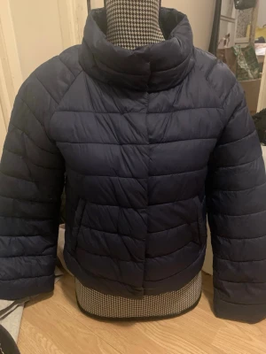 Mörkblå pufferjacka med hög krage - Säljer en mörkblå pufferjacka med hög krage och breda kanaler. Jackan har en boxig passform och stängs framtill. Perfekt för kalla dagar när du vill hålla dig varm och snygg. Materialet är syntet och jackan har två sidofickor. Har ett litet håll i fickan