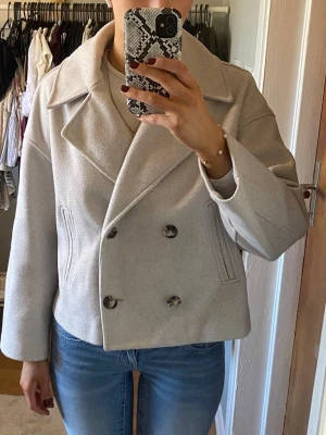 Beige kort rock med bred krage - Stilren beige rock med dubbelknäppning och bred krage. Modellen är kortare och har raka ärmar samt två snedställda fickor framtill. Perfekt för dig som gillar en klassisk men modern look. Materialet ser ut att vara ullblandning. Lite nopprig på vissa ställen. 