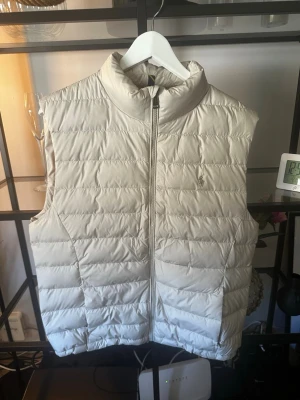  Ralph Lauren - Snygg beige dunväst från Ralph Lauren med quiltad design och dragkedja framtill. Västen har hög krage och två sidofickor, samt den klassiska lilla loggan broderad på bröstet. Perfekt för lager-på-lager och stilren look.