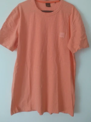 Persikofärgad t-shirt från BOSS - Persikofärgad t-shirt från BOSS med rund halsringning och korta ärmar. Materialet är mjukt och lätt bomull, perfekt för en avslappnad look. Diskret logo på vänster sida. Enkel och stilren design som är lätt att matcha.