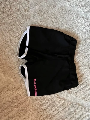 Svarta BLACKPINK shorts med vit kant passar barn och vuxna - Coola svarta shorts från BLACKPINK med vit kant runt benen och rosa logga på ena benet. De har en loose passform och är perfekta för träning eller chill. Materialet känns som polyester och de har elastisk midja för extra komfort.