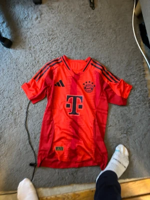 Bayern tröja - Tröjan är både snygg och stilren, ofta med vita detaljer som passar bra till det röda. Den är gjord av bra tyg som känns bekvämt och lätt, vilket gör att spelarna kan röra sig fritt under matcherna. Många fans tycker också att den är fin att ha på sig till vardags eftersom den ser både sportig och modern ut ⚽