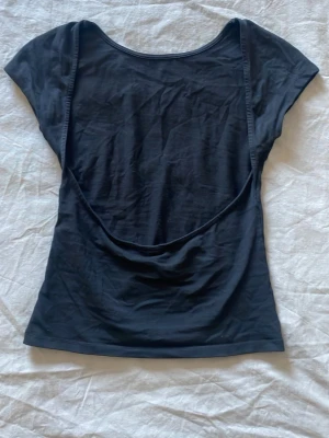 Svart topp med öppen rygg - Svart topp i bomull med korta ärmar och snyggt öppen rygg. Modellen är tight och har en enkel, stilren design som passar perfekt till jeans eller kjol. Perfekt för dig som gillar minimalistiska plagg med en twist. Obs ett litet hål under ena armen men inget som märks när man har den på sig 