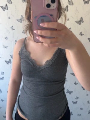 Grå ribbad linnetopp med spets - Snygg grå linnetopp med ribbad struktur och spetsdetaljer längs v-ringningen. Tunn och stretchig med smala axelband, perfekt för varma dagar eller som lager under en skjorta. Enkel att matcha till olika outfits. Kolla in min profil för fler fina och trendiga plagg!