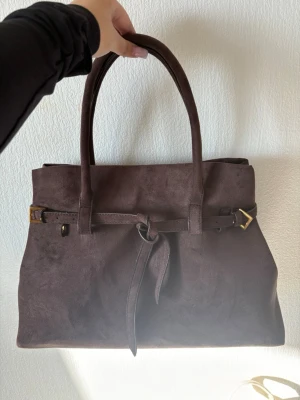 Mörkbrun mocka handväska - Stilren mörkbrun handväska i mjuk mocka med dubbla handtag och dekorativt bälte med guldfärgade detaljer framtill. Väskan har en klassisk, rektangulär form och är perfekt för dig som gillar en elegant look. Ingen synlig logga eller märke.