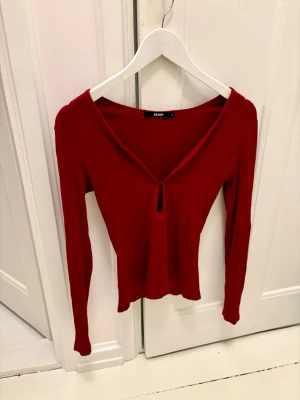 Superfin vinröd v-ringad topp från Bik Bok - Säljer en snygg röd långärmad topp från Bik Bok med djup v-ringning som går att knäppa. Toppen är i viskos vilket gör den väldigt skön på och lagom sval till varmare kvällar. Passformen är slim och den är enkel att matcha med jeans eller kjol.