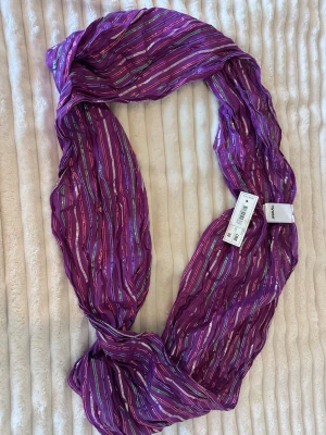 Lila halsduk med metallicränder mywear - Snygg lila halsduk från mywear med tunna metallicfärgade ränder i silver, grönt och rött. Halsduken är lätt och luftig, perfekt för att addera färg till din outfit. Materialet är tunt och något transparent, vilket ger en cool och trendig look.