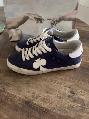 The gallerist sneakers - Snygga mörkblå sneakers i mocka från The gallerist med vita detaljer och vita skosnören. Skorna har en unik vit klöverdetalj på sidan och vit hälkappa. Klassisk låg modell med bekväm passform och slitstark sula. Knappt använda! 