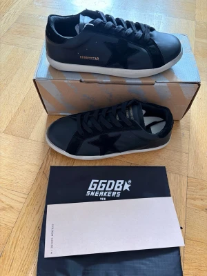Golden goose sneaker - Snygga svarta sneakers från Golden Goose med ikonisk stjärndetalj på sidan. Skorna har en vit sula, svarta skosnören och är tillverkade i en mix av läder och mocka. Perfekta för dig som gillar en clean och stilren look.