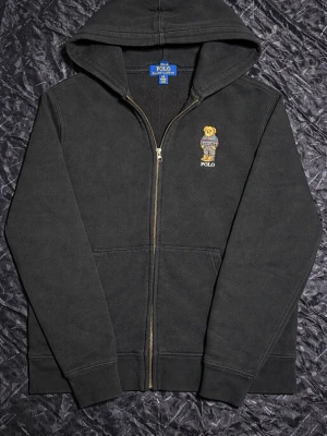 Svart hoodie från Polo Ralph Lauren - Svart hoodie med dragkedja från Polo Ralph Lauren. På bröstet finns den klassiska Polo Bear-broderingen. Hoodien har huva, två fickor fram och ribbade muddar. Perfekt för en avslappnad och stilren look.