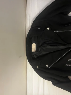 Svart bikerjacka från NLY Trend - Snygg svart bikerjacka från NLY Trend med silvriga dragkedjor och knappar. Jackan har klassisk krage och flera fickor med dragkedja. Perfekt för en cool och stilren look.