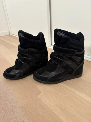 Trendiga skor - Snygga svarta sneakers från Buffalo med kilklack och högt skaft. Skorna har en mix av mocka och läderdetaljer samt tre breda kardborreband över foten. Perfekta för dig som vill ha en cool och unik look med extra höjd.