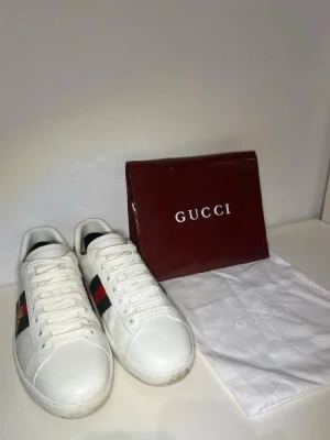Gucci Ace sneakers  - Snygga vita Gucci Ace sneakers i läder med klassiska gröna och röda sidoremsor samt broderad guldfärgad bi på sidan. Skorna har vit sula, vita skosnören och låg profil. Perfekta för dig som vill ha en clean och lyxig look.