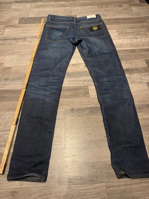 Mörkblå jeans från Stone Island - Säljer ett par mörkblå jeans från Stone Island med klassisk femficksmodell och raka ben. Jeansen har en snygg tvätt och den ikoniska Stone Island-loggan på bakfickan. Materialet är slitstark denim i bomull. Perfekta för dig som gillar streetwear och stilren design. Dom sitter som W28 L32