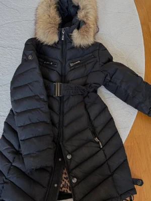 Svart dunjacka från Hollies - Snygg svart dunjacka från Hollies , bälte i midjan och flera dragkedjefickor. Jackan har quiltad design och detaljer i metall. Perfekt för kalla vinterdagar. Välanvänd