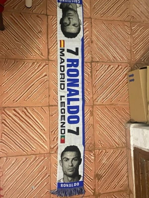 Vit och blå Ronaldo halsduk - Snygg vit och blå halsduk med texten '7 Ronaldo 7', 'Madrid Legend' och flaggor från Spanien och Portugal. Halsduken har porträtt av Ronaldo i svart och blått samt fransar i ändarna. Perfekt för dig som älskar fotboll och vill hylla en ikon.