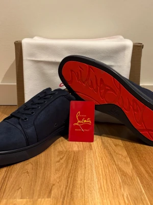 Marinblå sneakers från Christian Louboutin -  Blå sneakers från Christian Louboutin i mocka med klassisk röd sula. Skorna har lågt skaft, svarta skosnören och en stilren design som sticker ut med den ikoniska röda undersidan.