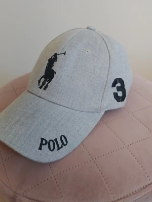 Grå keps från Polo Ralph Lauren - Snygg grå keps från Polo Ralph Lauren med klassisk svart polospelare broderad framtill, svart text 'POLO' på skärmen och en stor svart siffra 3 på sidan. Tillverkad i mjukt tyg med böjd skärm och justerbar passform.