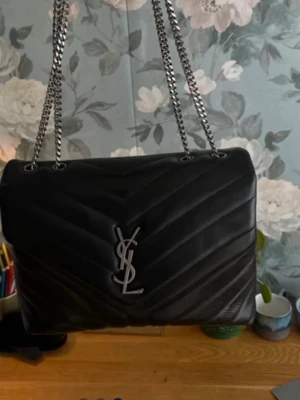 Svart YSL handväska i läder - Säljer en svart quiltad handväska från Saint Laurent med silverfärgad YSL-logga framtill och kedjeremmar i metall. Väskan är tillverkad i mjukt läder och har ett innerfack med dragkedja. Perfekt för dig som gillar lyxiga accessoarer.