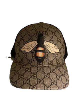 Gucci keps med bi-motiv och GG-mönster - Säljer en sällsynt äkta keps från Gucci. Framtill finns ett stort bi-motiv i svart, gult och vitt. Kepsen har böjd skärm och mesh på sidorna. Perfekt för dig som vill sticka ut med en exklusiv accessoar. Kepsen säljs inte längre och går prisad för över 5000kr
