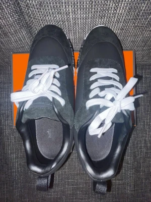 Svarta sneakers med vit sula - Snygga svarta sneakers med vit, räfflad sula och vita skosnören. Ovandelen är i en mix av mocka och textil, med detaljer i läderimitation. Skorna har en låg profil och breda hällor baktill för enkel påtagning. Perfekta för en trendig streetstyle-look.