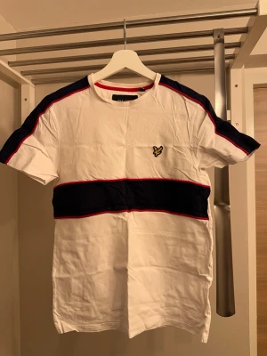 Vit t-shirt med blåa och röda detaljer - Snygg vit t-shirt från Lyle & Scott med breda marinblå ränder och röda detaljer över bröstet och axlarna. Klassisk passform och rund hals. Ikonisk gul logga broderad på bröstet. Perfekt för en stilren och sportig look.