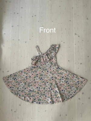 Blommig oneshoulder sommarklänning - Söt kort blommig klänning med volang och oneshoulder-design. Klänningen har ett brett axelband på ena sidan och volangdetalj över bröstet. Färgerna går i ljusa toner med inslag av rosa, grön och vit. Perfekt för varma dagar.