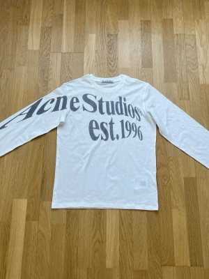 Vit långärmad t-shirt Acne Studios - Vit långärmad t-shirt från Acne Studios med stort svart tryck 'Acne Studios est. 1996' över bröstet och ena ärmen. Klassisk rund hals och rak passform. Perfekt för dig som gillar stilrena och trendiga plagg med statement-tryck.