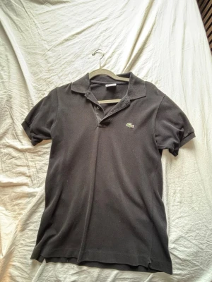 Svart pikétröja från Lacoste - Klassisk svart pikétröja från Lacoste den är i bra skick har 2 exakt likadan så om ni är intresserade kan ni få båda för ett schysst pris dom är både i storlek 2 och de motsvarar S