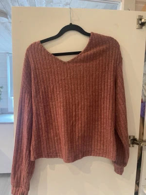 Rosa ribbstickad tröja med twist - Mysig rosa ribbstickad tröja med v-ringning fram och snygg twistad detalj i ryggen. Tröjan har långa ärmar och avslappnad passform, perfekt för en trendig och bekväm look. Materialet känns mjukt och stretchigt.