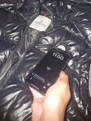 Svart dunväst från Moncler - Snygg svart dunväst från Moncler med glansig finish och klassisk Moncler-logga på bröstet. Västen har dragkedja framtill, två sidofickor med dragkedja och en hög krage med huva. Perfekt för lager-på-lager och streetwear-vibe.