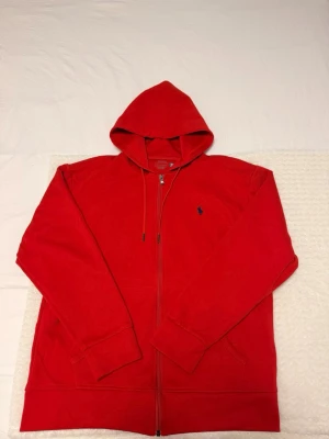 Röd hoodie från Polo Ralph Lauren - Snygg röd Polo Ralph Lauren hoodie i slim stil/passform som sitter snyggt på kroppen. Klassisk design med hel dragkedja, justerbar huva och den ikoniska broderade Polo-loggan på bröstet. Praktiska framfickor och mjukt, bekvämt material som passar perfekt både till vardags och en mer uppklädd casual look.