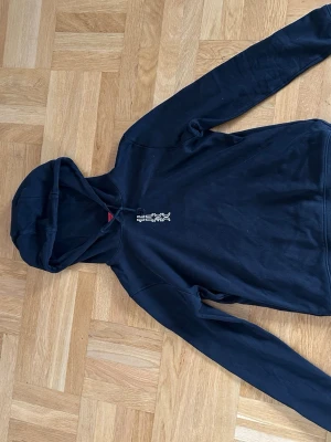 Svart HUGO hoodie med grafiskt tryck - Säljer en svart hoodie med huva och vitt grafiskt tryck både fram och bak. Hoodien har en klassisk passform och är tillverkad i mjukt bomullsmaterial. Perfekt för dig som gillar streetwear och vill ha en stilren men ändå unik look.
