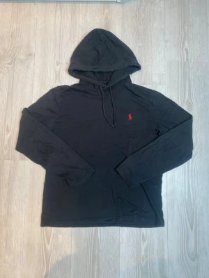 Svart hoodie från Polo Ralph Lauren - Säljer en svart hoodie från Polo Ralph Lauren med den klassiska röda loggan broderad på bröstet. Hoodien har dragsko i huvan och är tillverkad i mjukt bomullstyg. Perfekt för en avslappnad och stilren look.