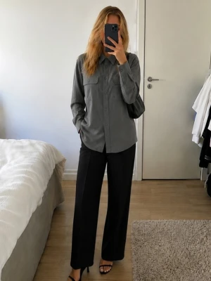 Mörkgrå skjorta med fickor - Stilren mörkgrå skjorta med klassisk krage och två bröstfickor. Skjortan har långa ärmar och knappar framtill. Perfekt för en clean och enkel look. Materialet ser ut att vara bomull eller polyester.