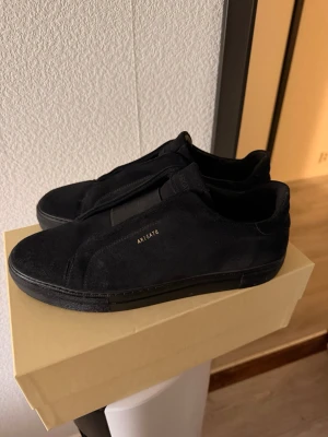 Svarta sneakers från Axel Arigato - Stilrena helsvarta sneakers från Axel Arigato i mocka med diskret guldfärgad logga på sidan. Lågt skaft och klassisk siluett med svart sula och ovandel. Perfekta för dig som gillar minimalistisk och clean stil. ÄKTA! Med kvitto 