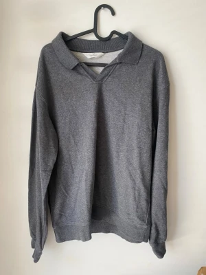 Grå college-tröja med krage H&M - Mysig grå college-tröja från H&M med klassisk krage och v-ringning. Tröjan har långa ärmar och ribbade muddar vid ärmslut och nederkant. Perfekt för en avslappnad och stilren look.