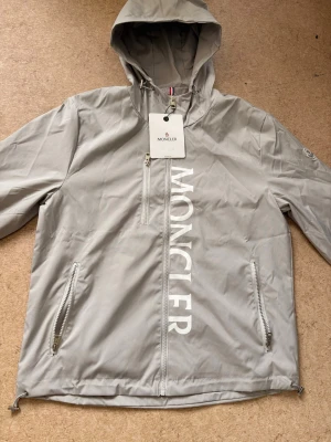 Ljusgrå vindjacka från Moncler - Snygg ljusgrå vindjacka från Moncler med huva och dragkedja framtill. Jackan har stora vita Moncler-bokstäver längs dragkedjan, två sidofickor med dragkedja och en bröstficka. Tillverkad i lätt, vattenavvisande material. Perfekt för en trendig streetwear-look.