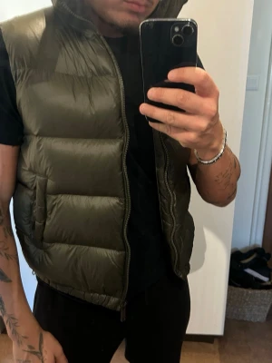 Olivgrön dunväst med hög krage - Säljer en olivgrön dunväst med hög krage och breda, vadderade paneler. Västen har dragkedja framtill och en clean, minimalistisk look. Perfekt att slänga över en hoodie eller t-shirt för extra värme. Märke och storlek saknas.