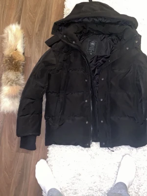 Svart dunjacka från Canada Goose - Svart dunjacka från Canada Goose med avtagbar huva och pälsdetalj. Jackan har quiltad design, ribbade muddar och logopatch på ärmen. Perfekt för kalla vinterdagar och har en klassisk, stilren look.