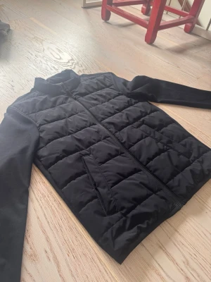 Svart pufferjacka med dragkedja - Säljer en stilren svart pufferjacka med raka ärmar och ståkrage. Jackan har quiltad design, två snedställda fickor framtill och stängs med dragkedja. Perfekt för kalla dagar och enkel att matcha till olika outfits.